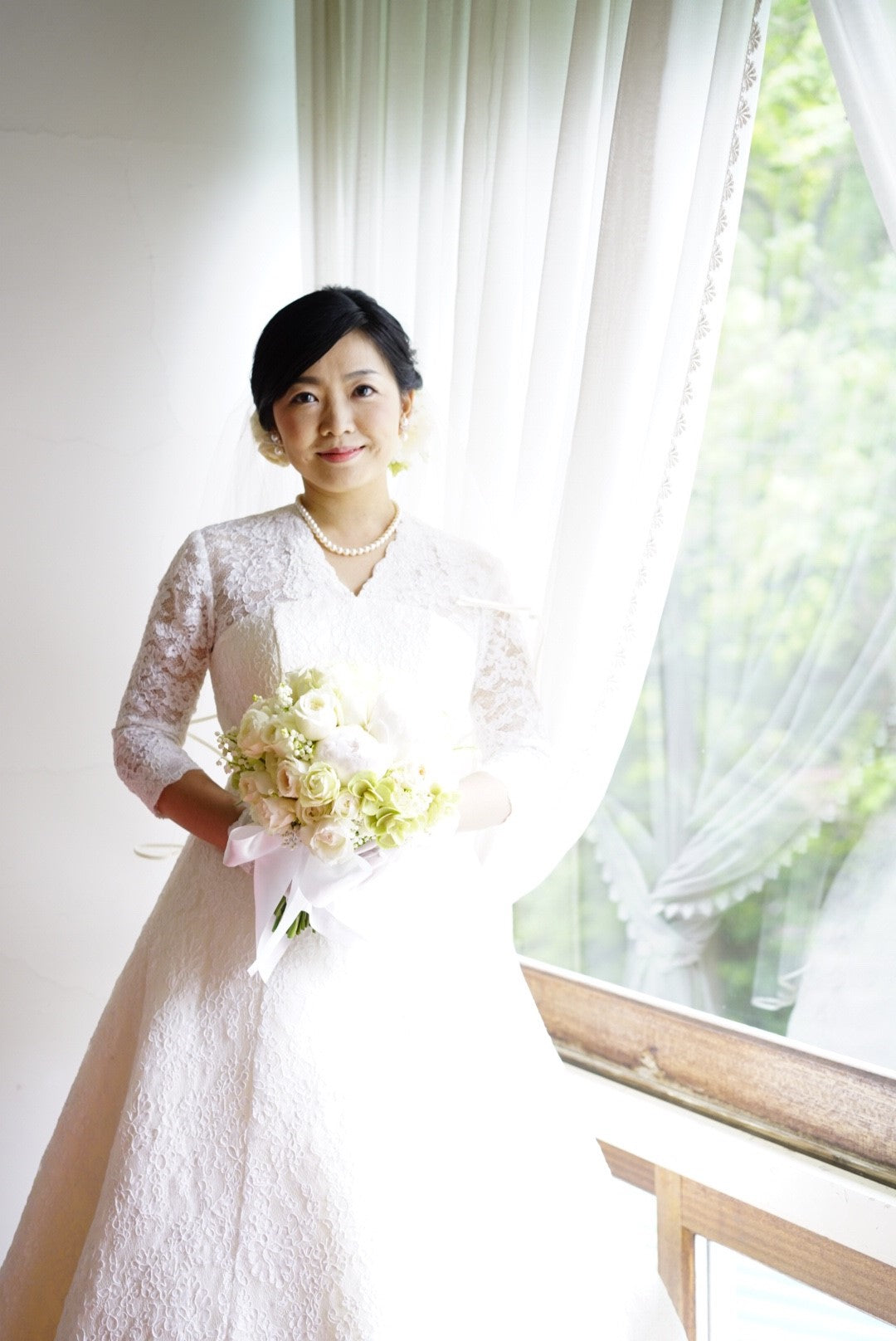 箱根・富士屋ホテルの洋装の花嫁様。白いレースの長袖ドレスとベール、パールのネックレス姿で窓辺に立ち、白とグリーン・淡いピーチピンクの芍薬・バラ・鈴蘭のクラッチブーケを持つ