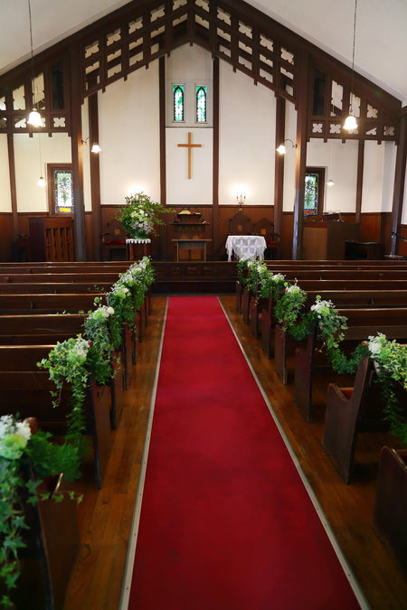 安藤記念教会の挙式装花の全景。赤いバージンロードと木造の祭壇・十字架、両脇にグリーンと白のチェアフラワーが並ぶ