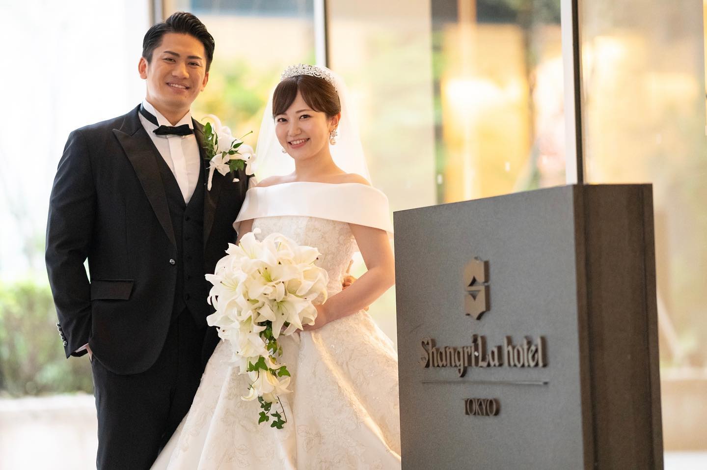 シャングリ・ラ 東京の入口サイン前で、カサブランカだけのキャスケードブーケを持つ花嫁様と、ユリのブートニアの新郎様｜一会アトリエ